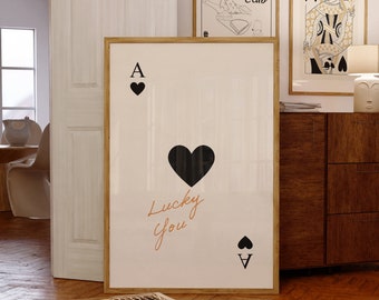 May include: A framed print of an ace of spades playing card with a black heart in the center and the text "Lucky You" written in orange. The card is on a white background.
