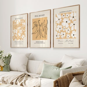 K&ouml;nnte beinhalten: Drei minimalistische Kunstdrucke mit beigefarbenem Hintergrund und wei&szlig;en Akzenten. Der linke Druck zeigt ein Blumenmuster und den Text "MATISSE" und "BERGGRUEN & CIE". Der mittlere Druck zeigt eine Linienzeichnung von T&auml;nzern und den Text "PICASSO" und "MUS&Eacute;E D'ORSAY". Der rechte Druck zeigt ein Blumenmuster und den Text "FLOWERS & PLANTS" und "Henri Matisse".