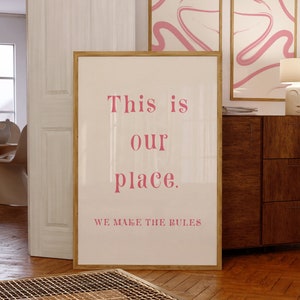 May include: A framed poster with the text "This is our place. We make the rules" in pink lettering. The poster is in a room with wooden floors and a wooden cabinet.
