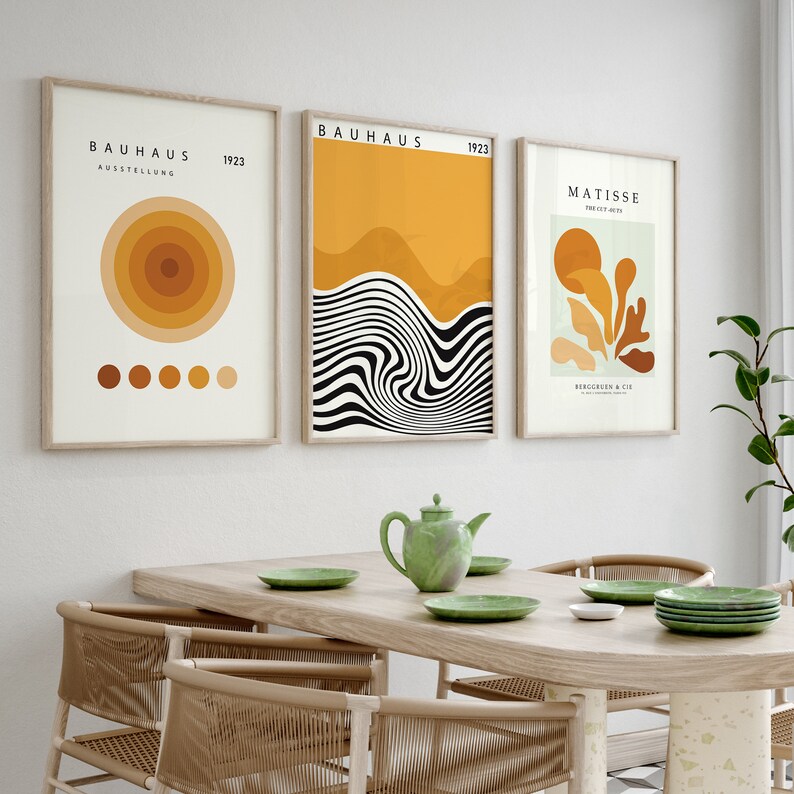 K&ouml;nnte beinhalten: Drei gerahmte Kunstdrucke mit abstrakten Designs in Gelb-, Orange- und Braunt&ouml;nen. Die Drucke h&auml;ngen an einer wei&szlig;en Wand &uuml;ber einem Holztisch mit gr&uuml;nen Tellern und einer gr&uuml;nen Teekanne.