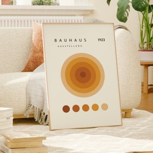 K&ouml;nnte beinhalten: Ein beiger Poster mit dem Text "BAUHAUS AUSSTELLUNG 1923" und einem gro&szlig;en konzentrischen Kreisdesign in Orange- und Braunt&ouml;nen. Am unteren Rand des Posters befinden sich sechs kleinere Kreise in einer Reihe.