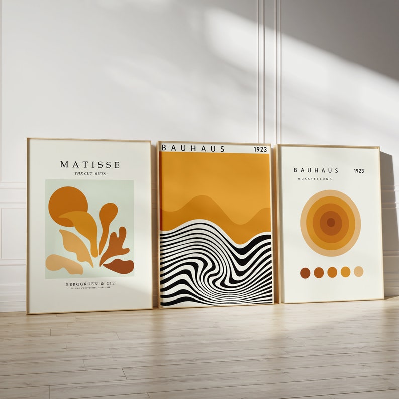 K&ouml;nnte beinhalten: Drei gerahmte Kunstdrucke mit minimalistischem Design. Der linke Druck zeigt ein von Matisse inspiriertes abstraktes Design in Orange-, Braun- und Gr&uuml;nt&ouml;nen mit dem Text "Matisse The Cut-Outs" und "Berggruen & Cie." Der mittlere Druck zeigt ein von Bauhaus inspiriertes abstraktes Design in Schwarz, Wei&szlig; und Gelb mit dem Text "Bauhaus 1923." Der rechte Druck zeigt ein von Bauhaus inspiriertes abstraktes Design in Orange- und Gelbt&ouml;nen mit dem Text "Bauhaus 1923 Ausstellung."