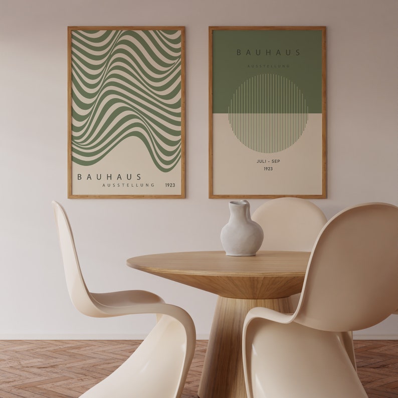 May include: Two framed posters with a green and white geometric design. The posters are titled "Bauhaus Ausstellung" and feature the dates "Juli - Sep 1923". The posters are hanging on a white wall above a wooden table with four white chairs.