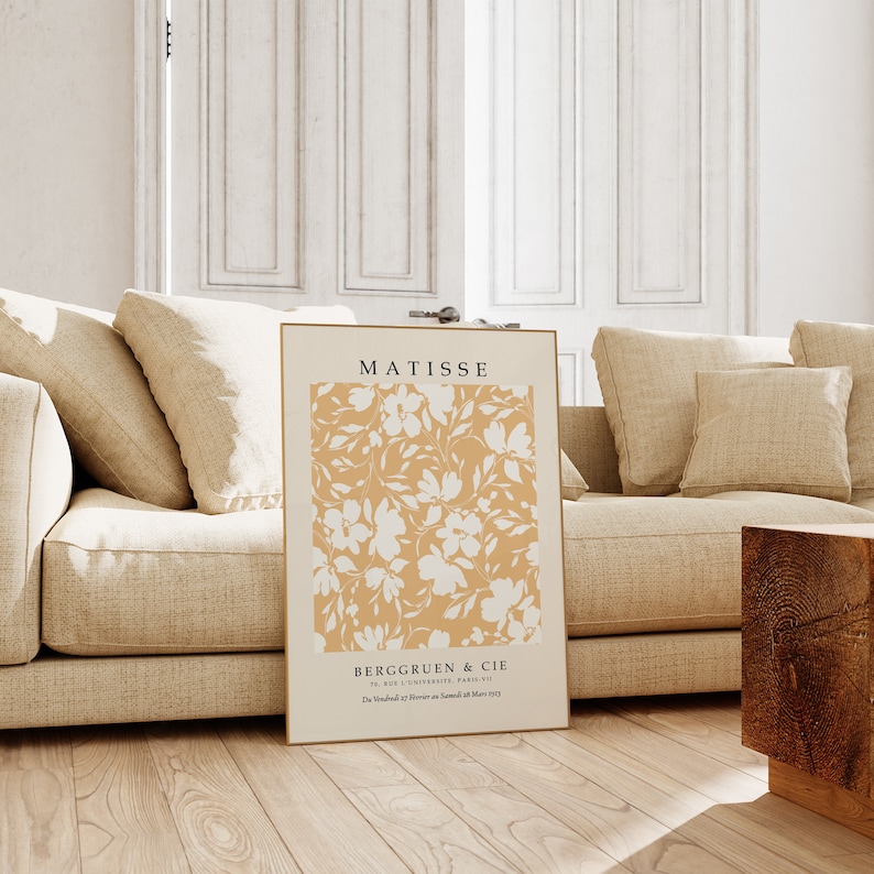 K&ouml;nnte beinhalten: Ein beige-wei&szlig;es Poster mit floralem Druck mit dem Text "MATISSE" und "BERGRUEN & CIE" in einem goldenen Rahmen. Das Poster lehnt an einem beigen Sofa mit Kissen.