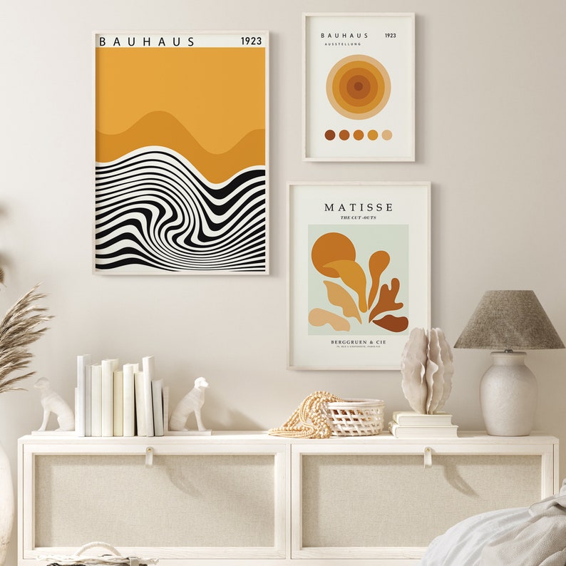 K&ouml;nnte beinhalten: Drei gerahmte Kunstdrucke mit abstrakten Designs in Schwarz, Wei&szlig; und Gelb. Die Drucke h&auml;ngen an einer wei&szlig;en Wand &uuml;ber einem wei&szlig;en Regal mit einer wei&szlig;en Lampe, B&uuml;chern und Dekoartikeln.