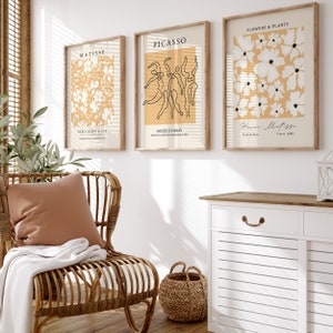 K&ouml;nnte beinhalten: Drei gerahmte Kunstdrucke mit minimalistischer &Auml;sthetik. Die Drucke zeigen Linienzeichnungen von Blumen, T&auml;nzern und abstrakten Formen auf einem hellgelben Hintergrund. Die Rahmen sind aus braunem Holz.