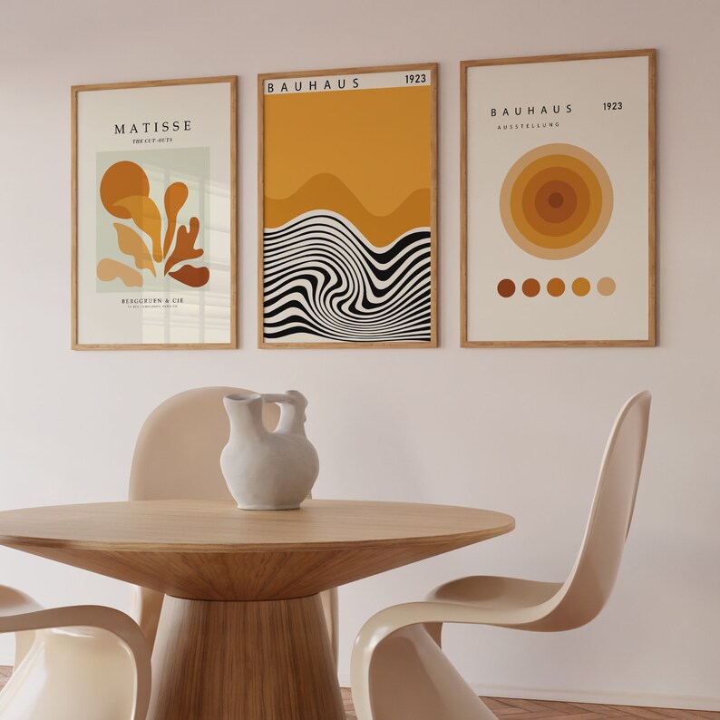 K&ouml;nnte beinhalten: Drei gerahmte Drucke mit abstrakten geometrischen Designs in Gelb-, Orange- und Braunt&ouml;nen. Die Drucke h&auml;ngen an einer hellrosa Wand &uuml;ber einem Holztisch mit zwei beigen St&uuml;hlen.