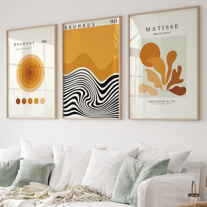 K&ouml;nnte beinhalten: Drei gerahmte Kunstdrucke mit minimalistischem Design. Die Drucke zeigen abstrakte Formen und Muster in Schwarz, Wei&szlig; und Gelb. Der obere Druck tr&auml;gt die Aufschrift "BAHAUS 1923" und zeigt ein schwarz-wei&szlig;es Wellenmuster. Der mittlere Druck tr&auml;gt die Aufschrift "BAHAUS 1923" und zeigt eine gelbe und orangefarbene abstrakte Form. Der untere Druck tr&auml;gt die Aufschrift "MATISSE THE CUT-OUTS" und zeigt einen gr&uuml;nen Hintergrund mit gelben und orangefarbenen abstrakten Formen.