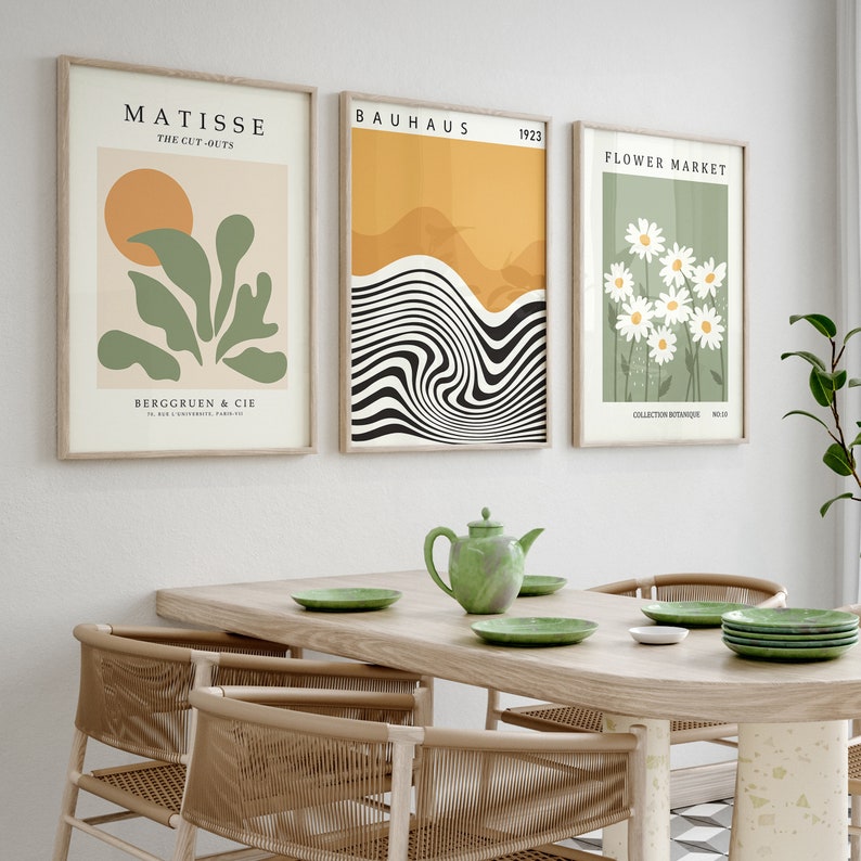 May include: Three framed art prints with minimalist designs. The left print features a green plant with a yellow sun in the background and the text "Matisse The Cut-Outs". The middle print features a black and white wavy design with a yellow and orange background and the text "Bauhaus 1923". The right print features white daisies on a green background with the text "Flower Market".