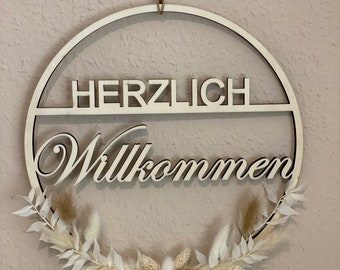 Op de afbeelding: Een houten cirkelkrans met de woorden "Herzlich Willkommen" in witte letters. De krans is versierd met gedroogd pampasgras en witte bladeren.