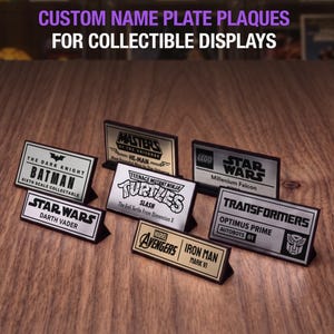 May include: A collection of rectangular name plate plaques for collectible displays, featuring various logos and character names including Batman, He-Man, Star Wars, Teenage Mutant Ninja Turtles, Transformers, and Avengers, with text reading CUSTOM NAME PLATE PLAQUES FOR COLLECTIBLE DISPLAYS and COLLECTOR SHELF.