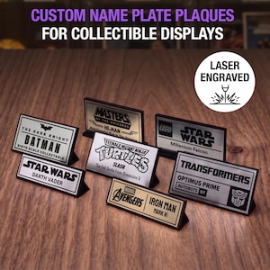 May include: A collection of rectangular custom name plate plaques for collectible displays, featuring laser-engraved text and logos for Batman, He-Man, Star Wars, Teenage Mutant Ninja Turtles, Transformers, and Marvel Avengers characters.
