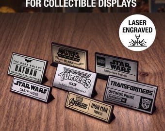 May include: A collection of rectangular custom name plate plaques for collectible displays, featuring laser-engraved text and logos for Batman, He-Man, Star Wars, Teenage Mutant Ninja Turtles, Transformers, and Marvel Avengers characters.