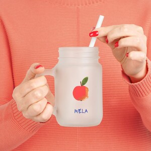 Peut inclure: Une tasse en verre givré avec une anse, représentant une pomme rouge avec une morsure et le mot "MELA" écrit en bleu. La tasse est tenue par une personne avec du vernis à ongles rouge.