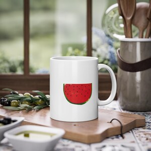 Op de afbeelding: Een witte keramische mok met een rode watermeloen-schijf afbeelding. De mok heeft een handvat en staat op een houten oppervlak.