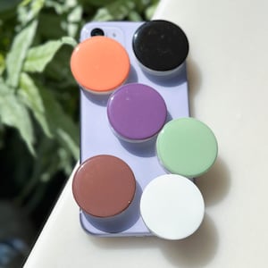 Puede incluir: Seis agarres para teléfono de diferentes colores, incluyendo naranja, morado, verde, marrón, blanco y negro. Los agarres están unidos a una funda de teléfono morada.