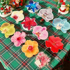 May include: A selection of flower-shaped coasters, including orchids and hibiscus, displayed on a green plaid surface. The coasters are in various colours such as yellow, pink, red, and orange. The text "GrassaCraft" is visible on the surface.