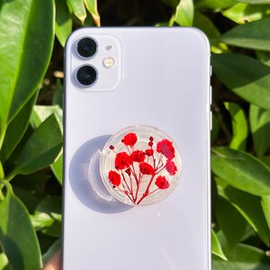 Peut inclure: Un support de téléphone transparent avec des fleurs rouges encapsulées dans de la résine, fixé à un smartphone blanc. La poignée est ronde et présente un motif floral. Le téléphone est placé sur un fond de feuilles vertes.