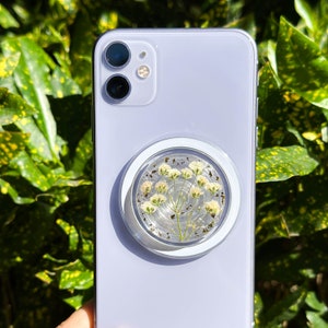 Puede incluir: Una funda de teléfono morada con un agarre de teléfono transparente con flores blancas prensadas.