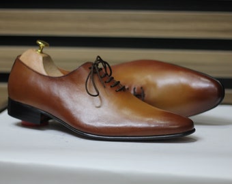 Zapatos Oxford de una sola pieza con acabado en pátina color coñac, suela de cuero bruñido a mano y cierre de cordones para hombre.