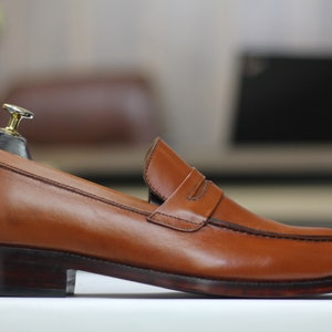 Puede incluir: Un mocas&iacute;n de cuero marr&oacute;n con una sola correa sobre el empeine. El mocas&iacute;n tiene una suela de cuero marr&oacute;n oscuro y una punta ligeramente redondeada.