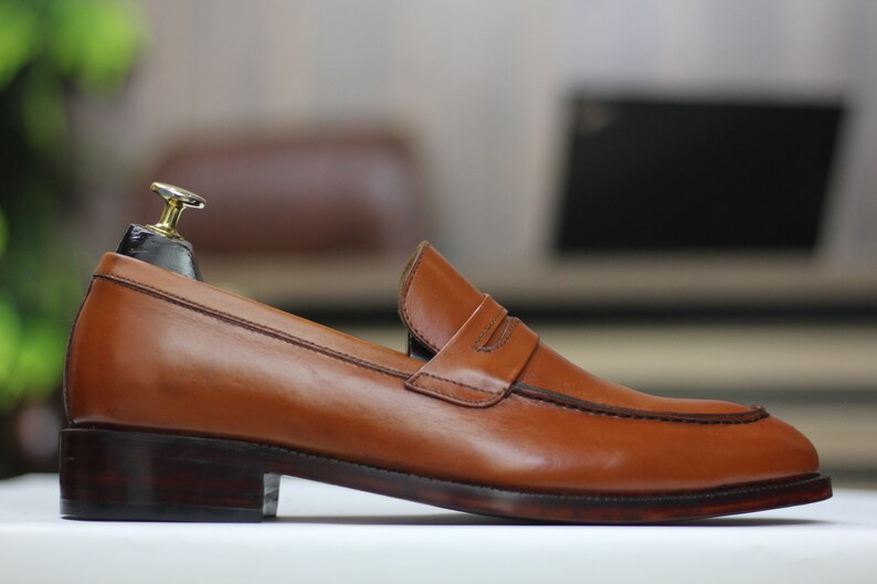 Puede incluir: Un mocas&iacute;n de cuero marr&oacute;n con una sola correa sobre el empeine. El mocas&iacute;n tiene una suela de cuero marr&oacute;n oscuro y una punta ligeramente redondeada.