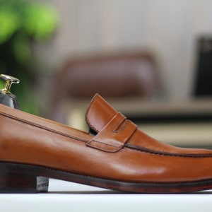 Puede incluir: Un par de mocasines de cuero marr&oacute;n con un detalle de tira penny. Los zapatos tienen una suela marr&oacute;n oscura y una punta ligeramente puntiaguda.