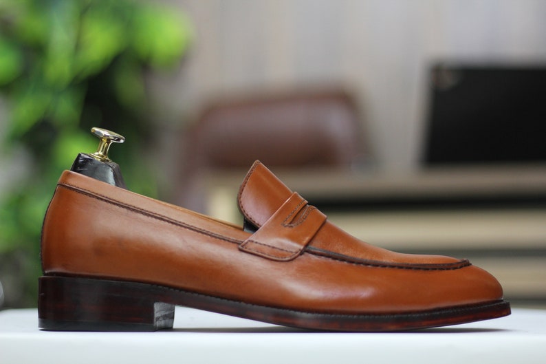 Puede incluir: Un par de mocasines de cuero marr&oacute;n con un detalle de tira penny. Los zapatos tienen una suela marr&oacute;n oscura y una punta ligeramente puntiaguda.