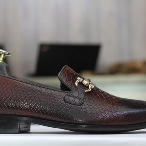 Puede incluir: Un mocas&iacute;n de cuero marr&oacute;n con estampado de piel de serpiente y una hebilla de metal dorado. El mocas&iacute;n tiene una punta puntiaguda y una suela de goma negra.