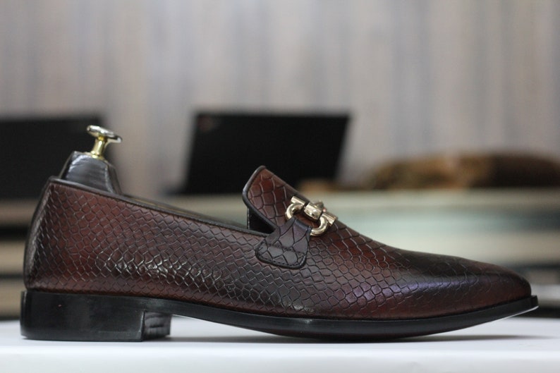 Puede incluir: Un mocas&iacute;n de cuero marr&oacute;n con estampado de piel de serpiente y una hebilla de metal dorado. El mocas&iacute;n tiene una punta puntiaguda y una suela de goma negra.