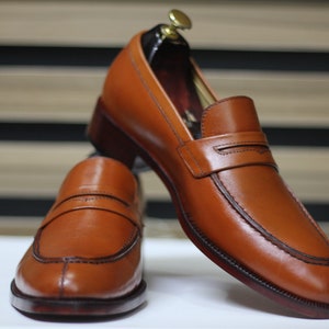 Puede incluir: Un par de mocasines de cuero marr&oacute;n con un detalle cosido en la punta y una sola correa en la parte superior. Los mocasines est&aacute;n sobre una superficie blanca.