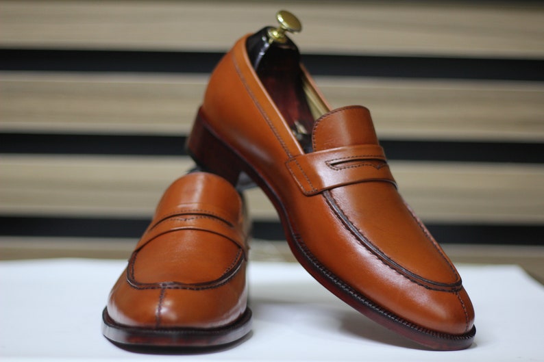 Puede incluir: Un par de mocasines de cuero marr&oacute;n con un detalle cosido en la punta y una sola correa en la parte superior. Los mocasines est&aacute;n sobre una superficie blanca.