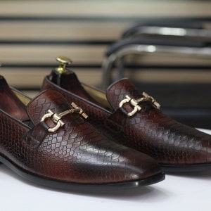 Op de afbeelding: Een paar bruine leren loafers met gouden gespen. De loafers hebben een getextureerd, slangachtig patroon.