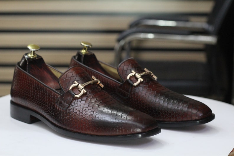 Puede incluir: Un par de mocasines de cuero marr&oacute;n con hebillas doradas. Los mocasines tienen un patr&oacute;n texturizado, similar a la piel de serpiente.
