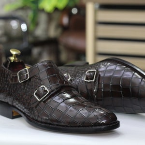 May include: A pair of brown leather monk strap shoes with a crocodile pattern. The shoes have two buckles on each strap.