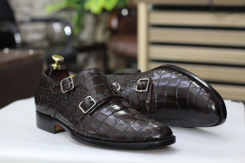 May include: A pair of brown leather monk strap shoes with a crocodile pattern. The shoes have two buckles on each strap.