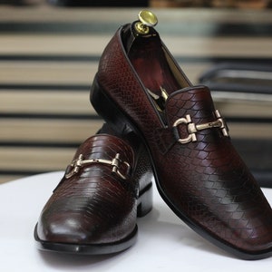 Puede incluir: Dos mocasines de cuero marr&oacute;n con hebillas doradas. Los mocasines tienen un patr&oacute;n texturizado de piel de serpiente.