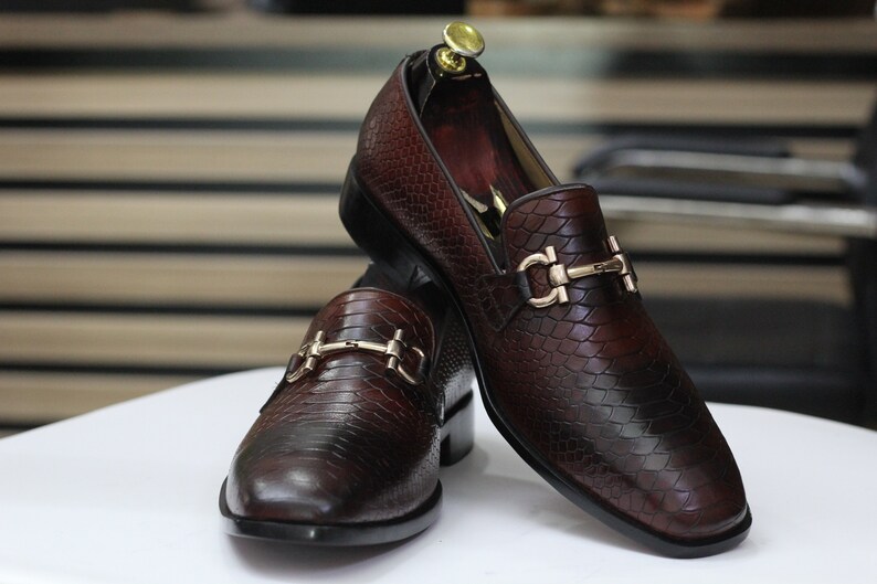 Puede incluir: Dos mocasines de cuero marr&oacute;n con hebillas doradas. Los mocasines tienen un patr&oacute;n texturizado de piel de serpiente.