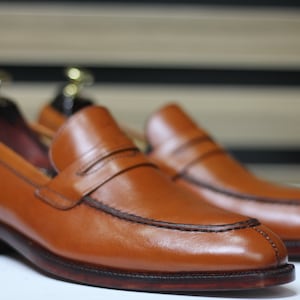 Puede incluir: Un par de mocasines de cuero marr&oacute;n con un detalle de tira penny. Los mocasines tienen una suela marr&oacute;n oscura y una punta ligeramente puntiaguda.