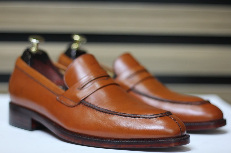 Puede incluir: Un par de mocasines de cuero marr&oacute;n con un detalle de tira penny. Los mocasines tienen una suela marr&oacute;n oscura y una punta ligeramente puntiaguda.