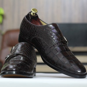 May include: A pair of brown leather monk strap shoes with a crocodile pattern. The shoes have a buckle closure and a pointed toe.