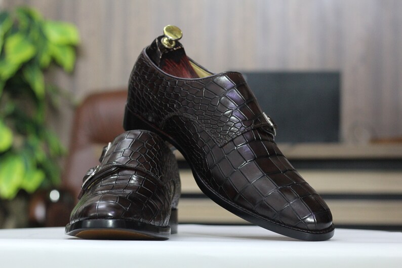 May include: A pair of brown leather monk strap shoes with a crocodile pattern. The shoes have a buckle closure and a pointed toe.