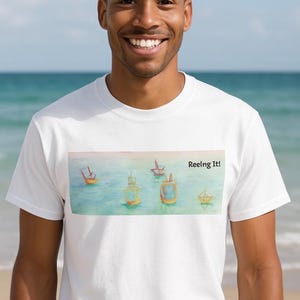 May include: White t-shirt featuring a watercolor painting of small boats on the water with the text "Reeling It!". The shirt is worn by a person smiling in front of a beach background.