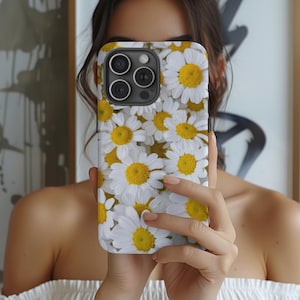 May include: A white iPhone case with a floral design featuring white daisies with yellow centers. The case has a camera lens cutout.