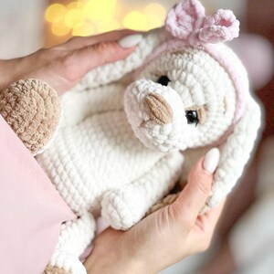 Pu&ograve; includere: Un peluche bianco lavorato a uncinetto con dettagli marroni, che indossa una fascia rosa con un fiocco. L'animale ha un'espressione dolce ed &egrave; tenuto da una mano.