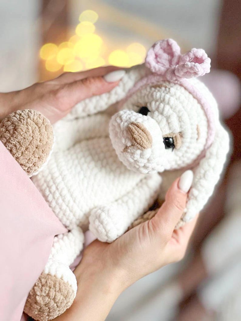 Pu&ograve; includere: Un peluche bianco lavorato a uncinetto con dettagli marroni, che indossa una fascia rosa con un fiocco. L'animale ha un'espressione dolce ed &egrave; tenuto da una mano.