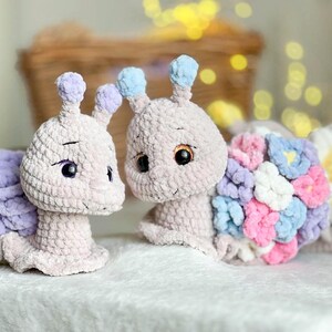 Op de afbeelding: Twee gehaakte slakken knuffels, een met een paarse schelp en de andere met een schelp gemaakt van kleurrijke gehaakte bloemen. De slakken zijn beide lichtroze en hebben zwarte ogen.