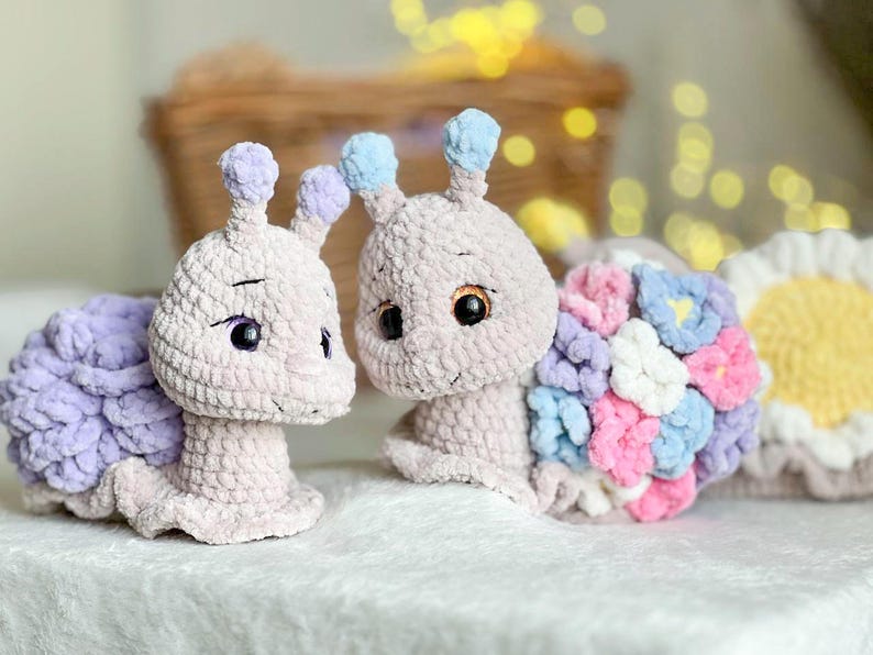 Op de afbeelding: Twee gehaakte slakken knuffels, een met een paarse schelp en de andere met een schelp gemaakt van kleurrijke gehaakte bloemen. De slakken zijn beide lichtroze en hebben zwarte ogen.