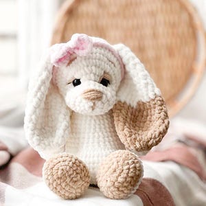 Pu&ograve; includere: Un giocattolo di peluche coniglio lavorato a uncinetto con pelliccia bianca e marrone. Il coniglio ha un fiocco rosa sulla testa e occhi di feltro marroni. Il coniglio &egrave; seduto su una coperta bianca e rosa.