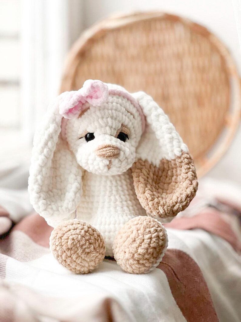 Pu&ograve; includere: Un giocattolo di peluche coniglio lavorato a uncinetto con pelliccia bianca e marrone. Il coniglio ha un fiocco rosa sulla testa e occhi di feltro marroni. Il coniglio &egrave; seduto su una coperta bianca e rosa.